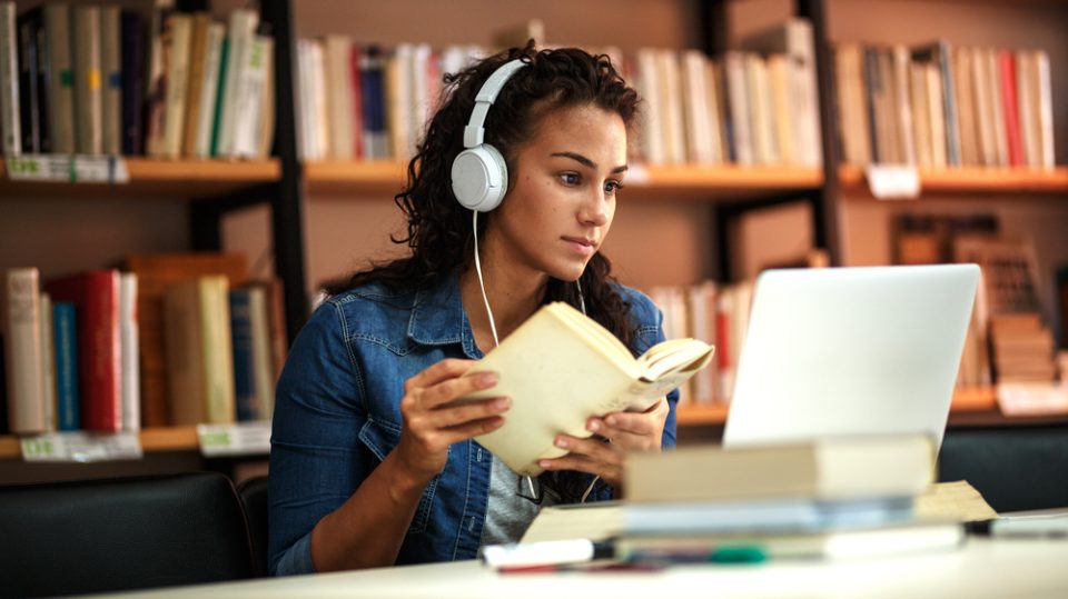 Woman with headphones over her head, holding a book and looking at an open laptop.