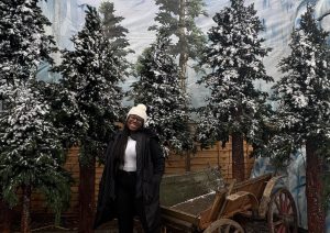 Woman standing in front of Christmas pine trees with a sprinkling of snow on them.